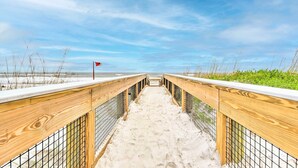 A wooden boardwalk leading to a beach with a red flag, surrounded by sand and sparse vegetation. - Modern Jacksonville Beach Retreat w/ Hot Tub! (Jacksonville Beach)