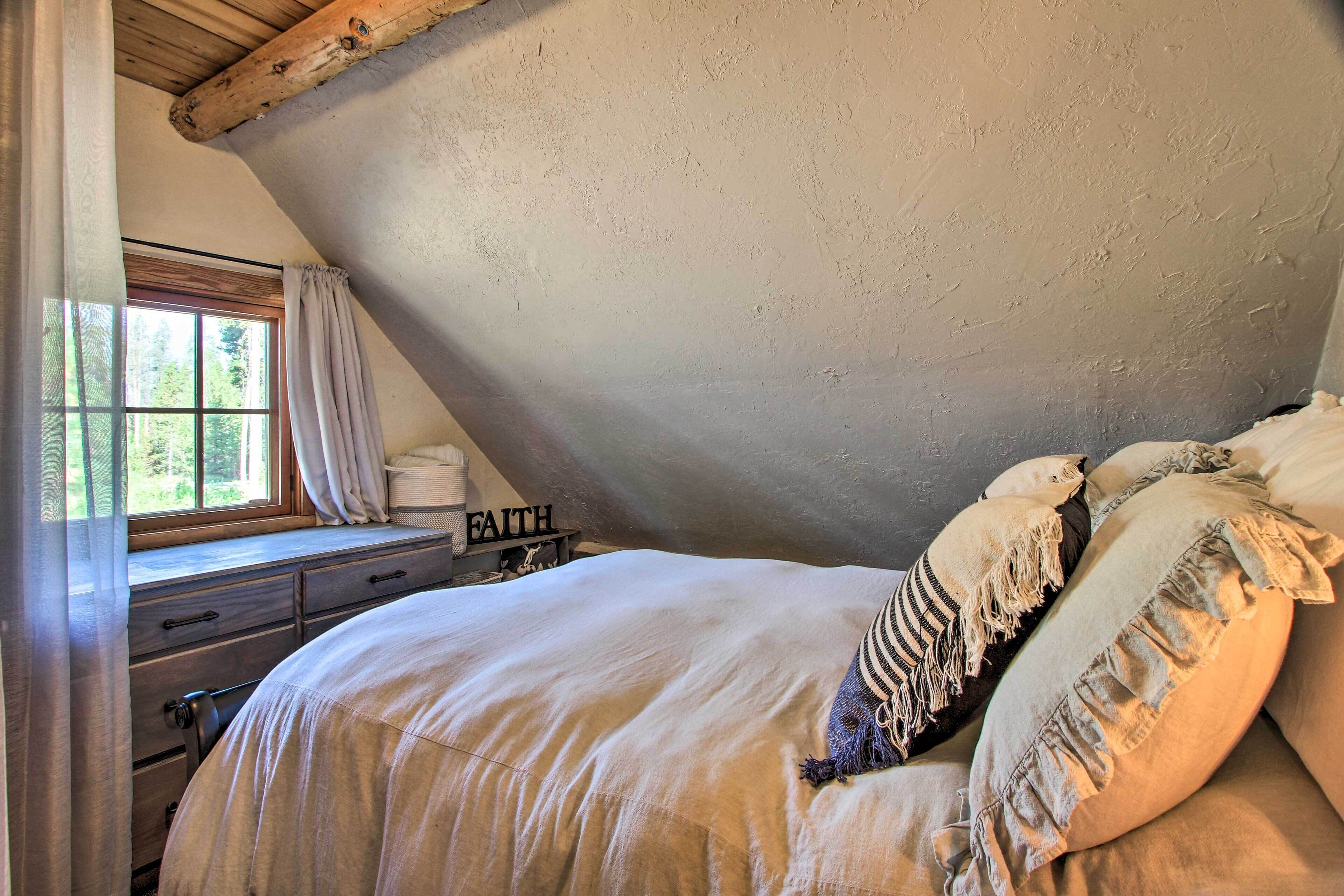 A cozy bedroom with a sloped ceiling, a large bed with pillows, a window with curtains, and a sign that reads "FAITH".