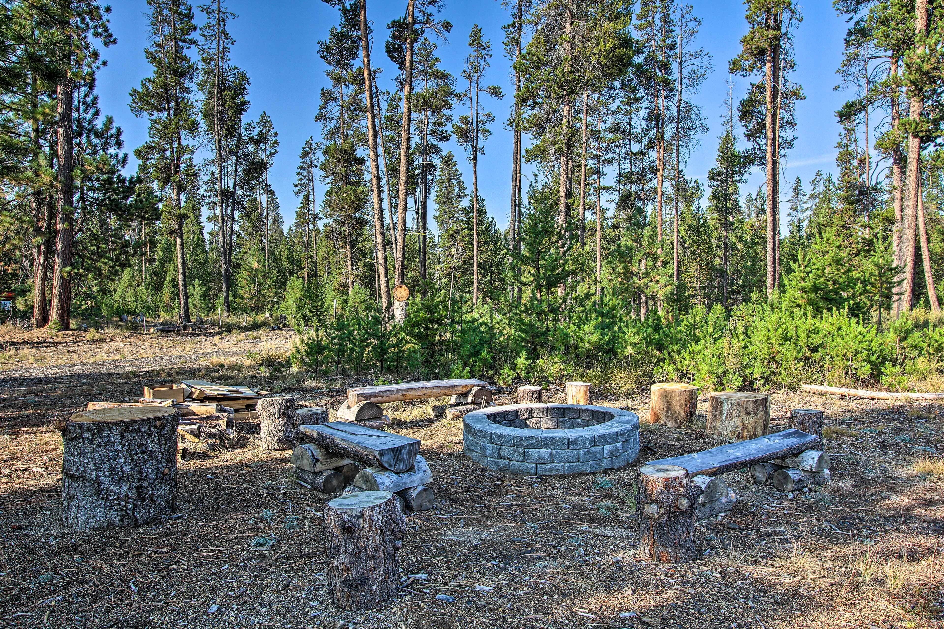 A forest clearing with a stone fire pit and benches, surrounded by tall pine trees.