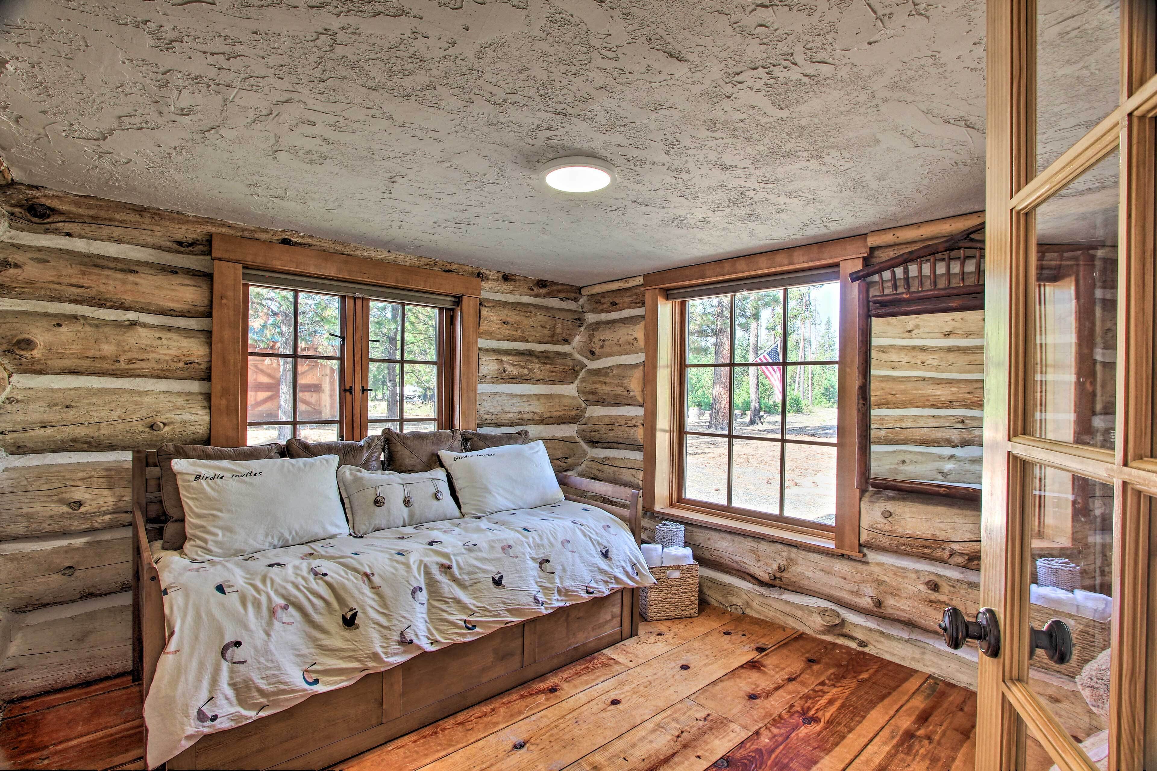 A log cabin bedroom with a bed, two windows, and a wooden ceiling.