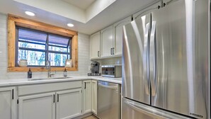 A modern kitchen with stainless steel appliances, white cabinetry, and a window with a view of trees. - Deck, Games & Fire Pit: Modern Pine Cabin! (Pine)