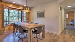 A dining area with a wooden table and chairs, a hanging light fixture, and a window with a view of trees. - Deck, Games & Fire Pit: Modern Pine Cabin! (Pine)