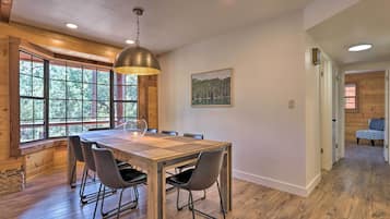 A dining area with a wooden table and chairs, a hanging light fixture, and a window with a view of trees.