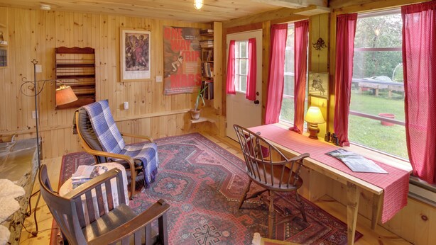 L’intérieur d’un chalet accueillant, avec un décor en bois, un bureau recouvert d’une nappe rouge, une chaise, une bibliothèque et une fenêtre ornée de rideaux rouges.