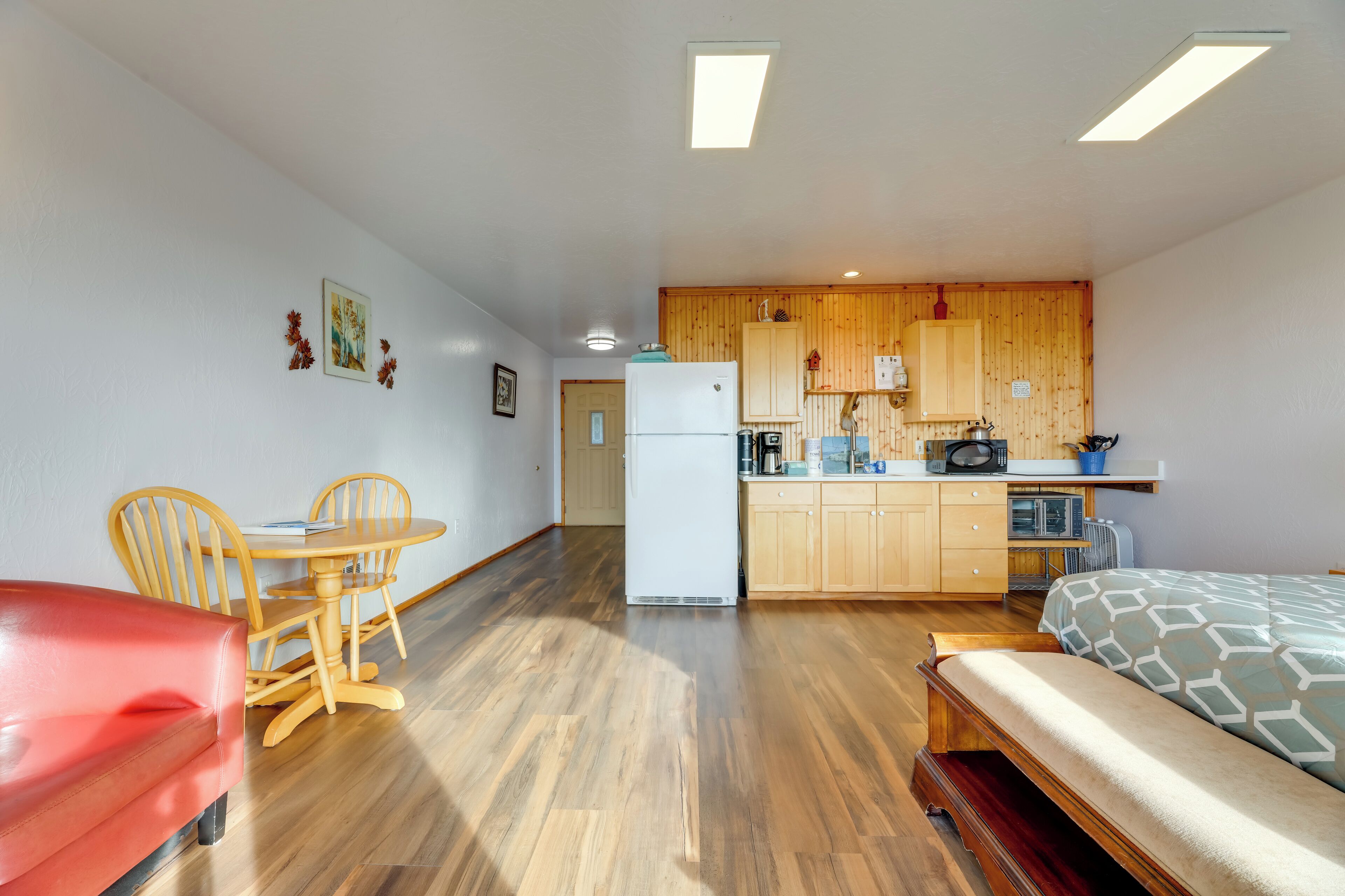 A compact room with a kitchenette, a dining area with wooden chairs, a bed with patterned bedding, and a red sofa.