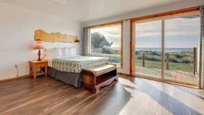 A bedroom with a large window, wooden flooring, a bed with a patterned bedspread, a nightstand with a lamp, and a view of the beach. - Sanderling Sea Cottages, Unit 14 (Waldport)