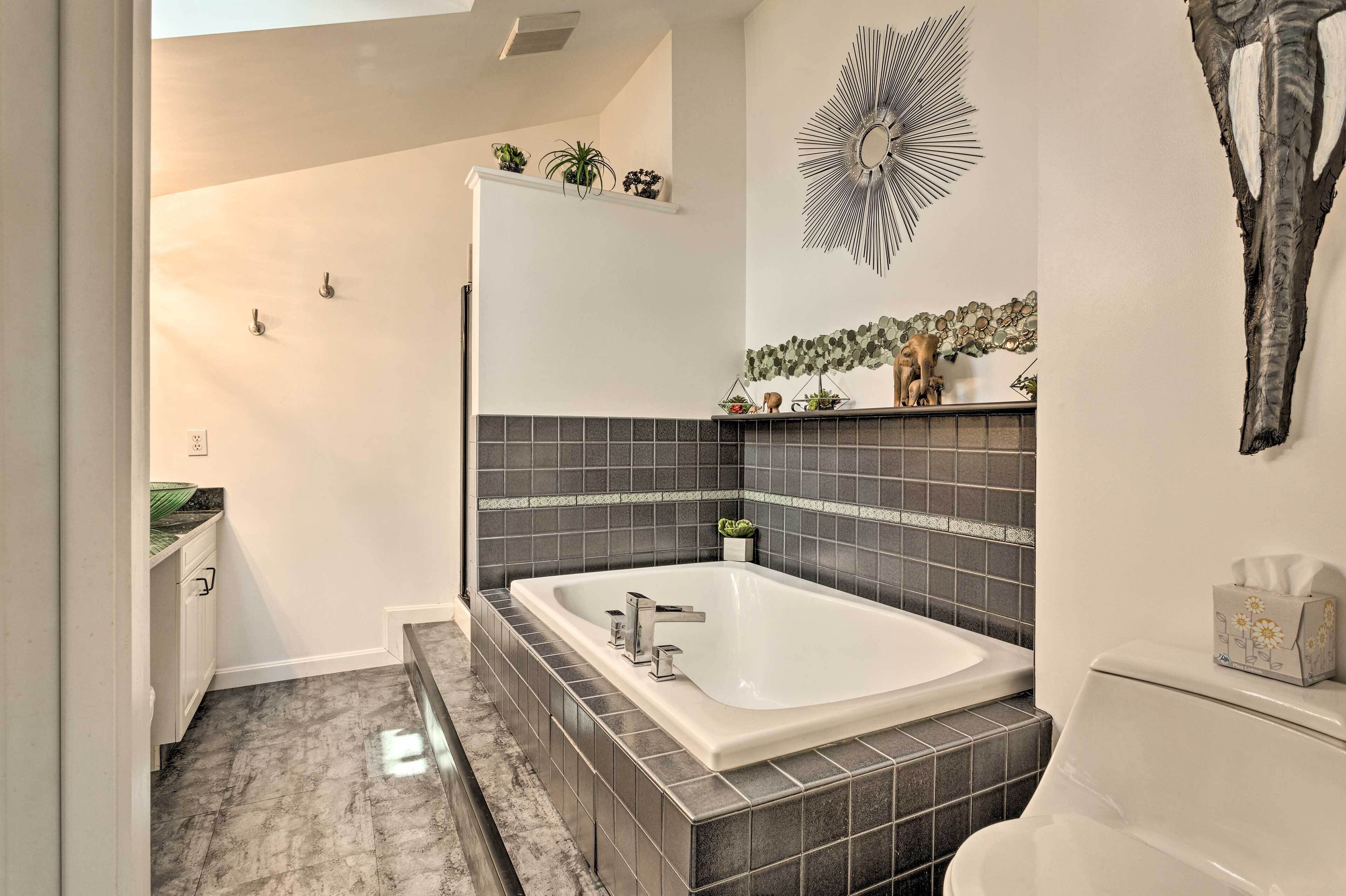 Une salle de bain avec une grande baignoire, des murs recouverts de carreaux foncés, un lavabo blanc et un miroir décoratif.