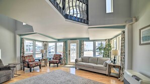 A spacious living room with a high ceiling, a staircase, and large windows offering a view of the outside. - Sun-soaked Coastal Cottage w/ Deck + Walk to Beach (Old Orchard Beach)
