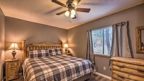 Une chambre à coucher comprenant un lit, deux tables de chevet avec des lampes, une commode en bois, un ventilateur de plafond et une fenêtre avec des rideaux.