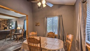 Un coin repas avec une table ronde recouverte d’une nappe, entourée de chaises en bois. Un ventilateur de plafond est suspendu au-dessus de la table, et il y a une fenêtre avec des stores.
