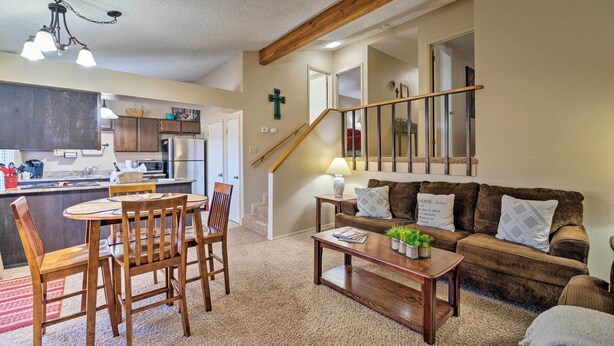 A cozy living room with a wooden coffee table, a brown sofa, and a dining area with wooden chairs and a table. A staircase with a wooden railing leads to another room. - Condo on Golf Course: 2 Mi to Grindstone Lake! (Ruidoso)