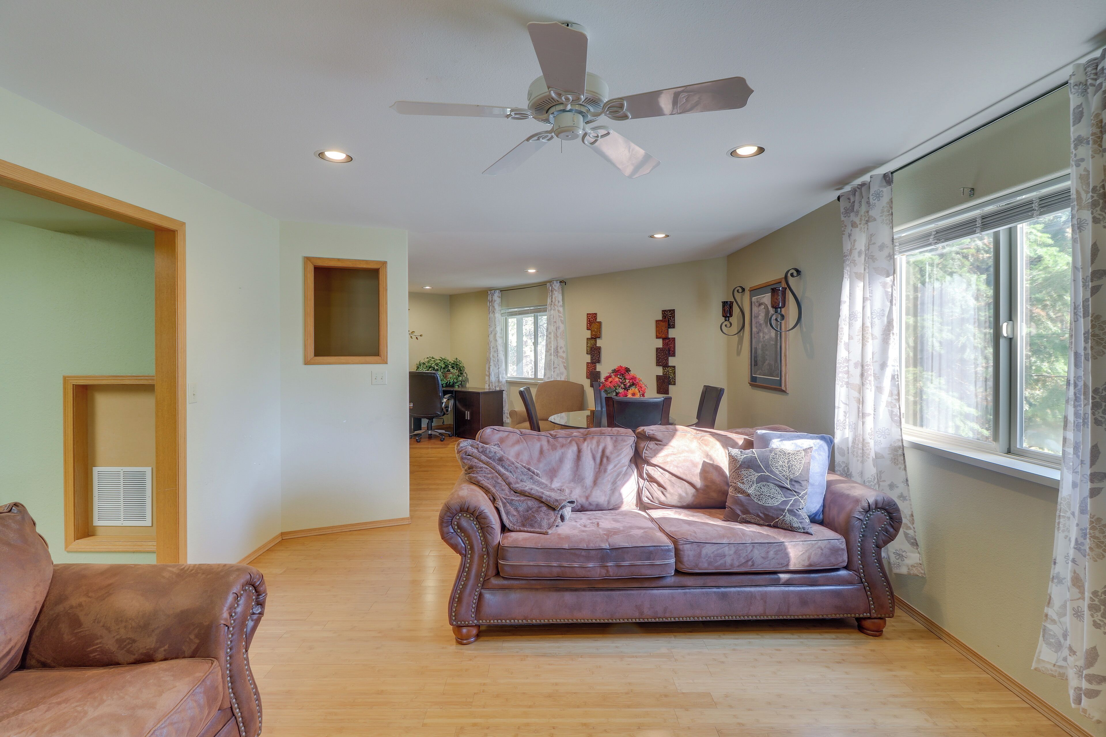 A spacious living room with a brown leather sofa, a wooden floor, a ceiling fan, and a large window with curtains.