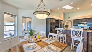 A dining area with a wooden table set for four, surrounded by white chairs, a chandelier, and a view of the kitchen area.