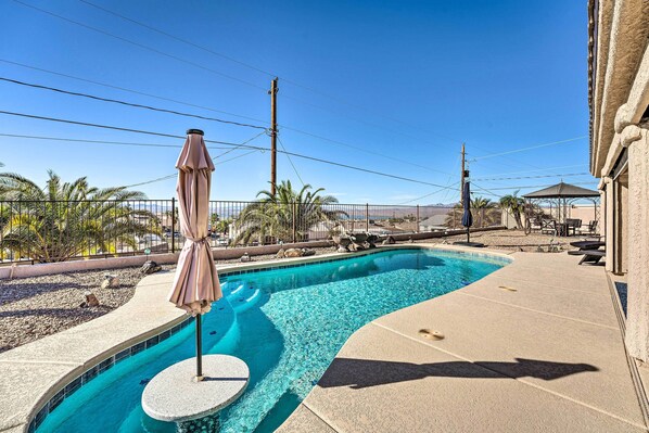 A swimming pool with a curved edge, surrounded by a concrete deck and a fence. There are palm trees and a gazebo in the background.