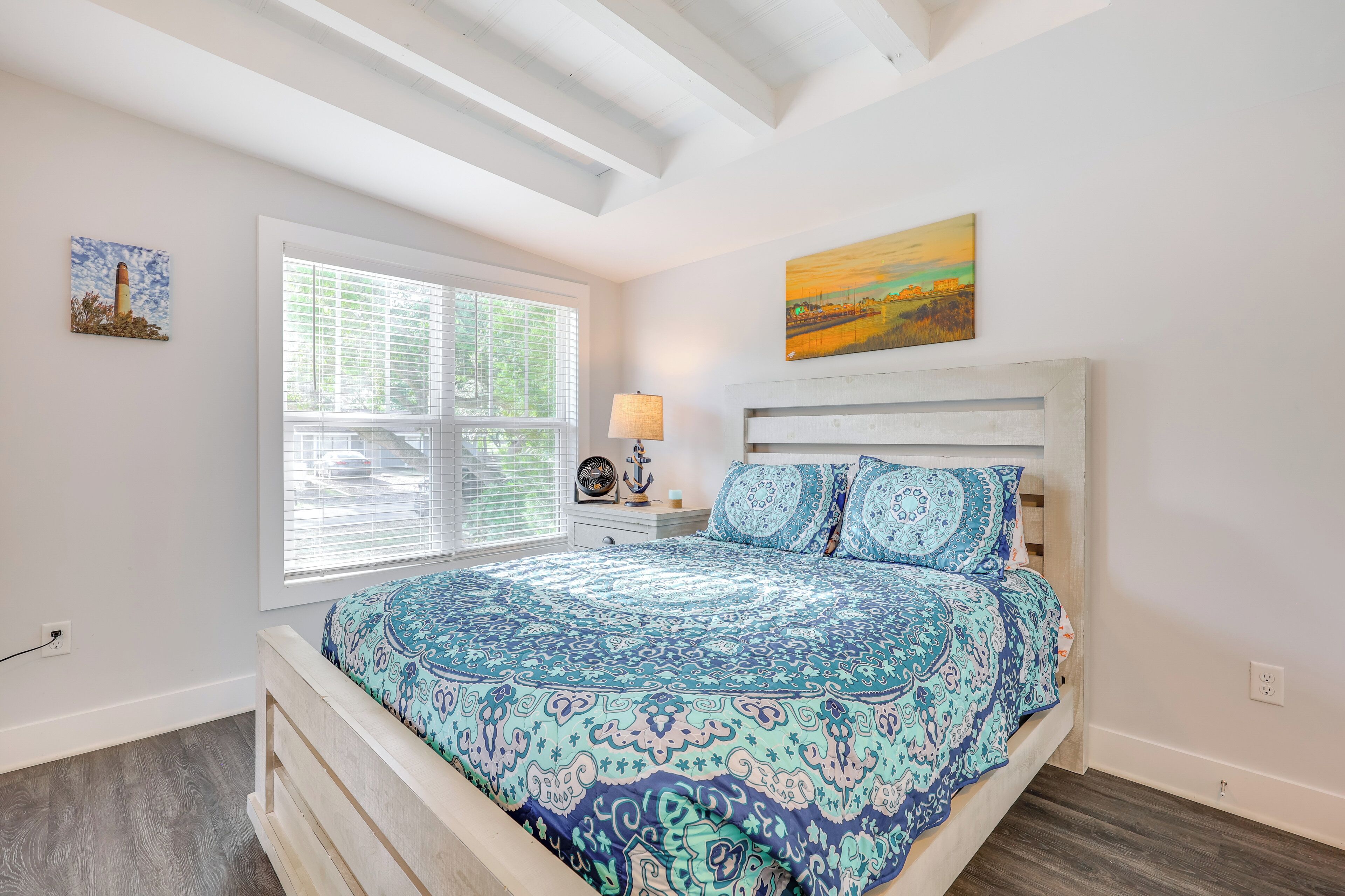 A bedroom with a large bed, a window with blinds, a nightstand with a lamp, and two wall-mounted paintings.