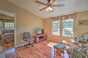 Une pièce avec un plancher en bois, un ventilateur de plafond, un téléviseur posé sur un meuble en bois et une fenêtre donnant sur l’extérieur.