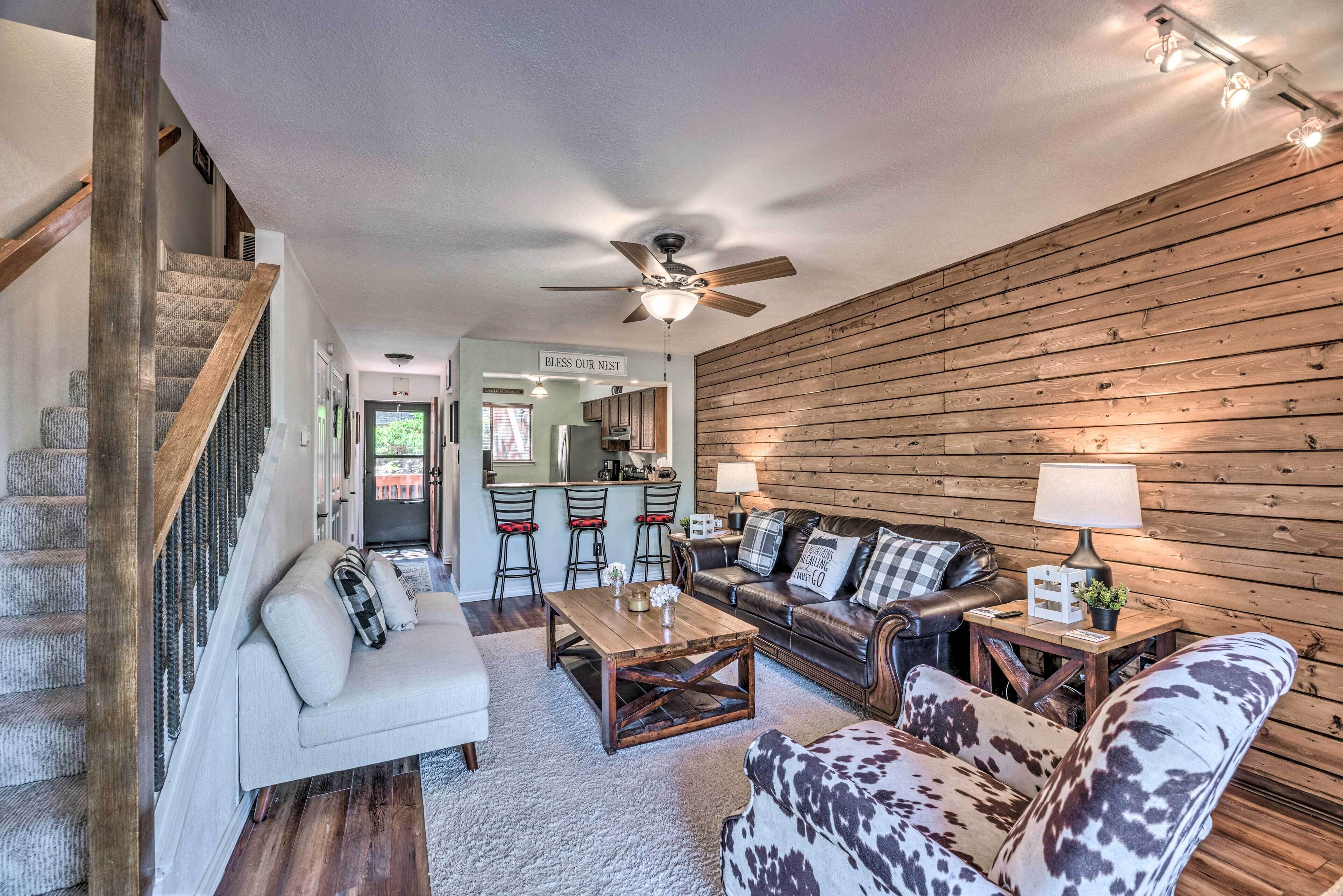 A spacious living room with a high ceiling, a wooden wall, a sofa set, a coffee table, and a dining area with a kitchen in the background.