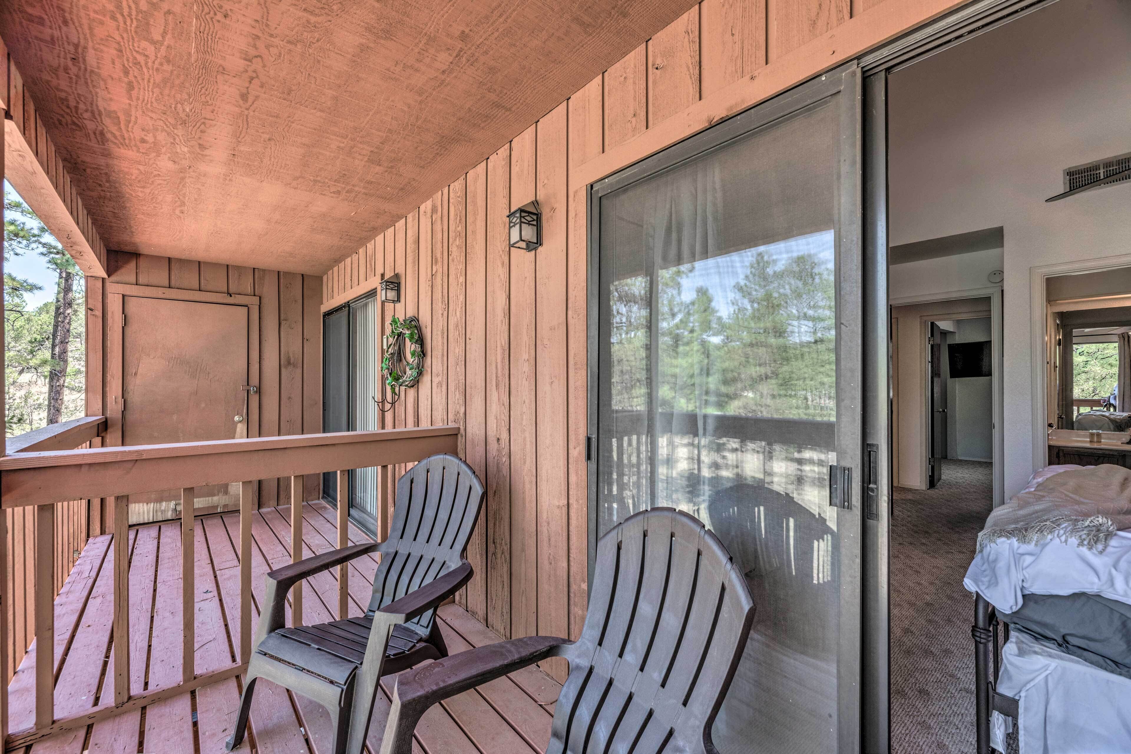 A wooden deck with Adirondack chairs, a sliding glass door leading to a bedroom, and a small wall-mounted light fixture.