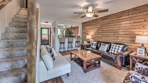 A living room with a staircase, a wooden wall, a ceiling fan, a sofa, a coffee table, and a kitchen area in the background. - Updated Ruidoso Mountain Escape w/ Deck & Balcony! (Ruidoso)