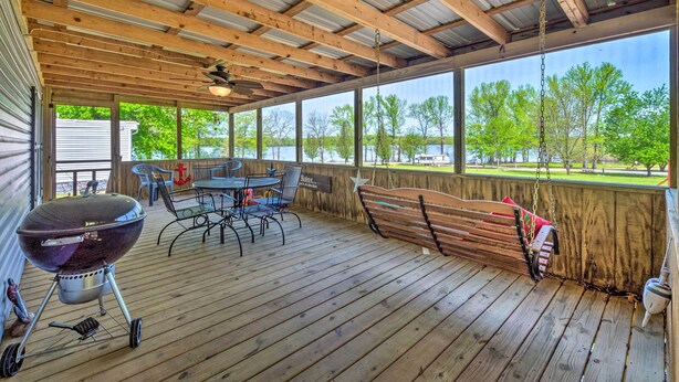 Un espacio al aire libre techado con terraza de madera, una parrilla, mobiliario exterior y vistas a un lago y árboles.