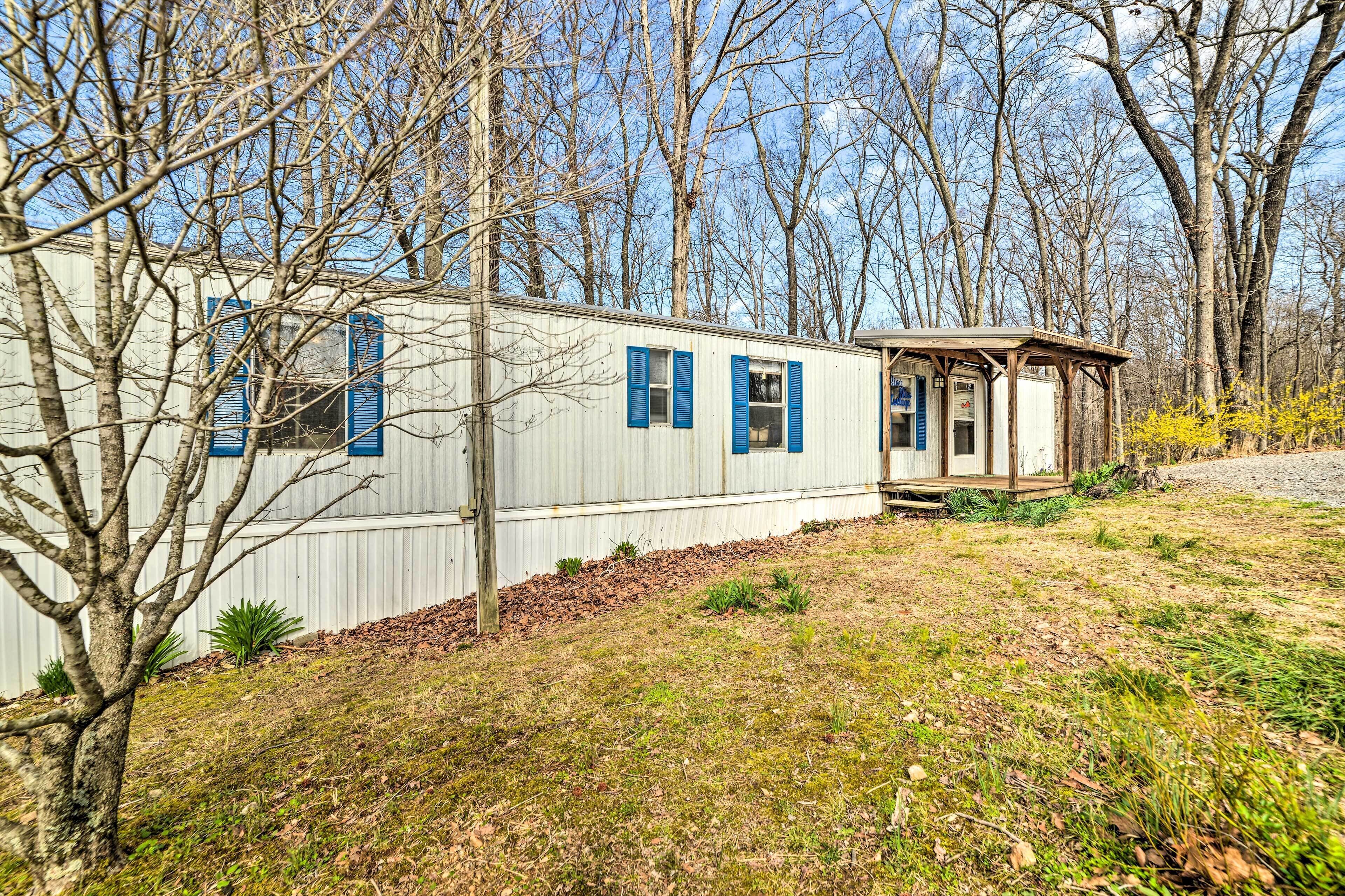 A mobile home with a small porch and a garage, surrounded by trees and a grassy area.