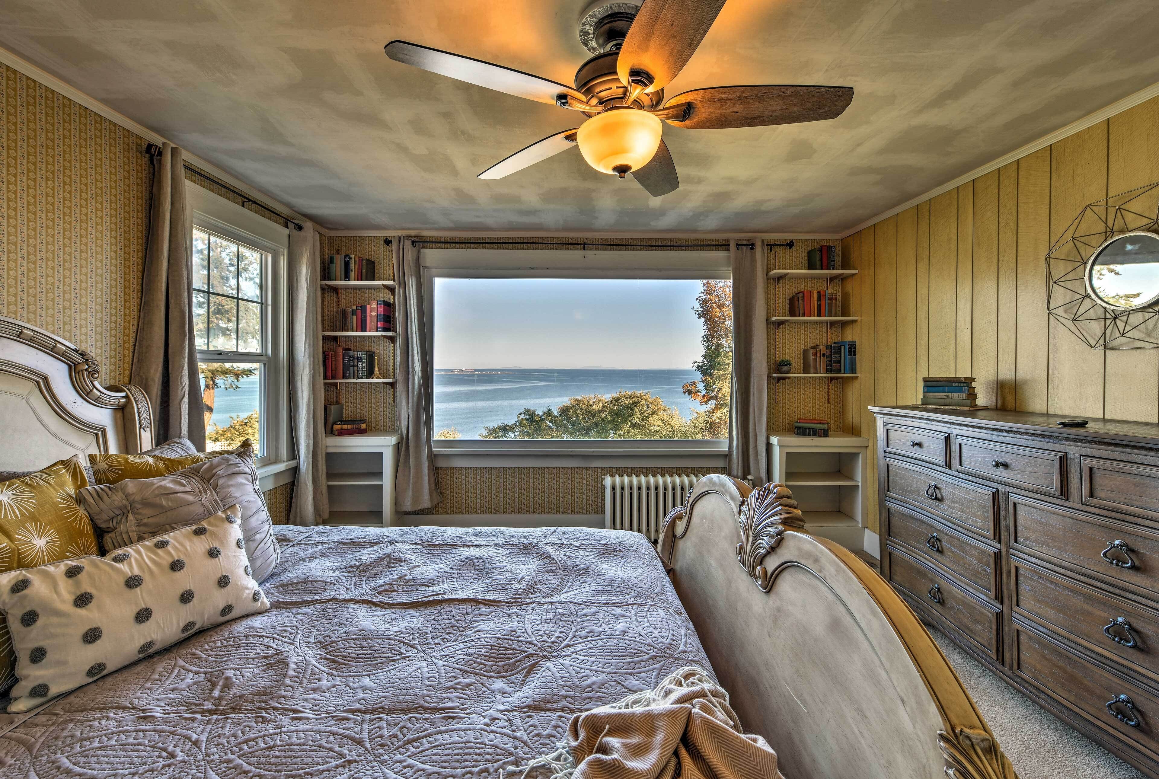 A bedroom with a large bed, a ceiling fan, a view of the sea through a window, and a bookshelf.