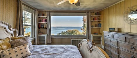 A bedroom with a large bed, a ceiling fan, a view of the sea through a window, and a bookshelf.