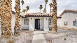 Une maison avec un toit en tuiles, un chemin en béton menant à l’entrée et des palmiers qui entourent la propriété.