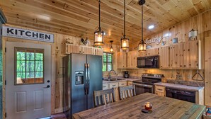 A wooden cabin kitchen with a stainless steel refrigerator, wooden cabinets, a dining table with chairs, and hanging pendant lights.