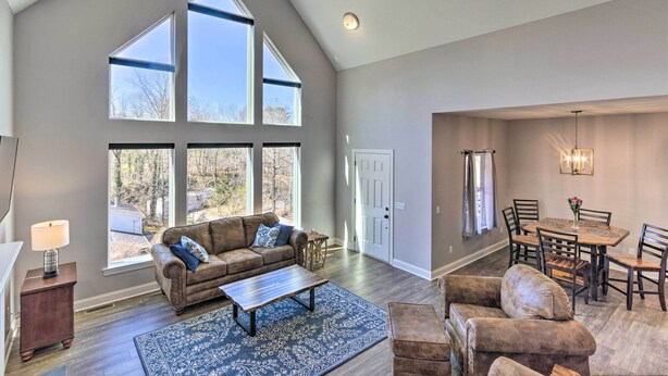 A spacious living room with a high ceiling, large windows, a brown leather sofa, a coffee table, and a dining area with a wooden table and chairs. - Family Home w/ Views ~ 5 Mi to Downtown Asheville! (Asheville)