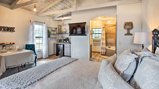 Un dormitorio espacioso con una cama grande, una zona de estar con una mesa y sillas, una kitchenette y un baño visible a través de un vano abierto.