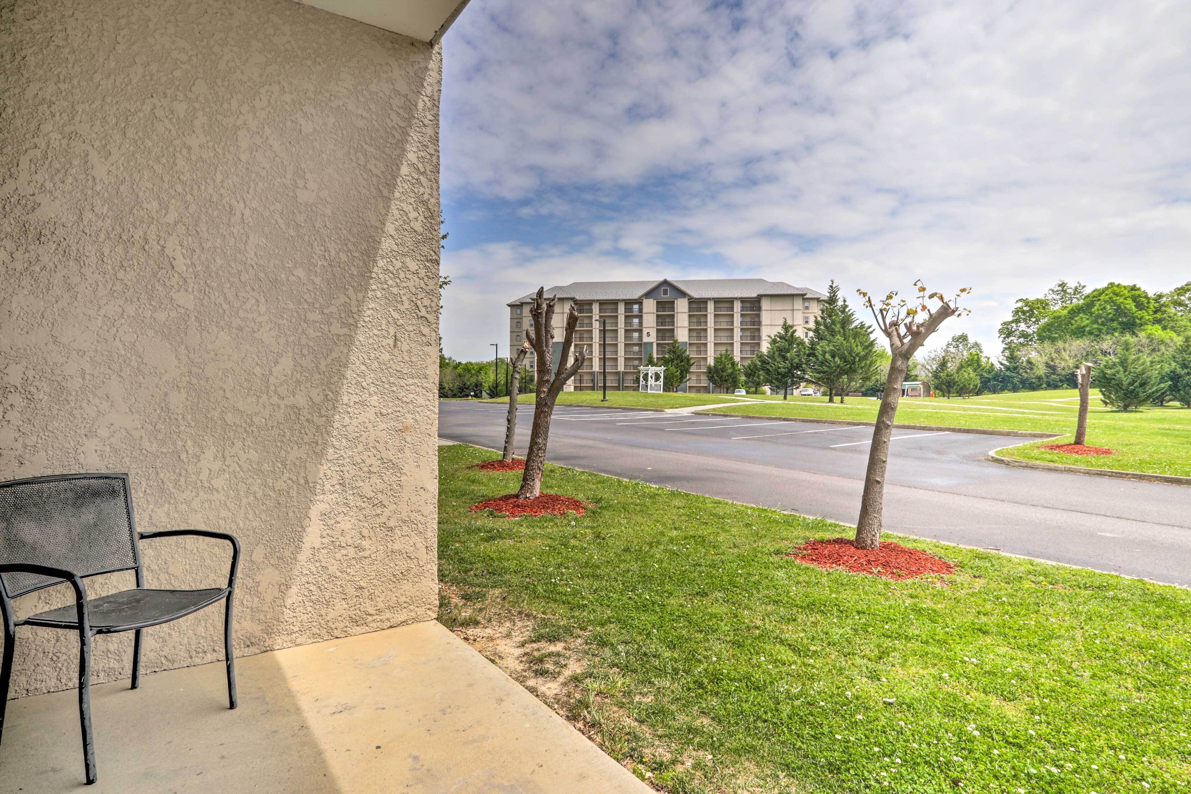 A building with a flat roof and multiple windows, a paved area with a few trees, and a chair on a concrete ledge.
