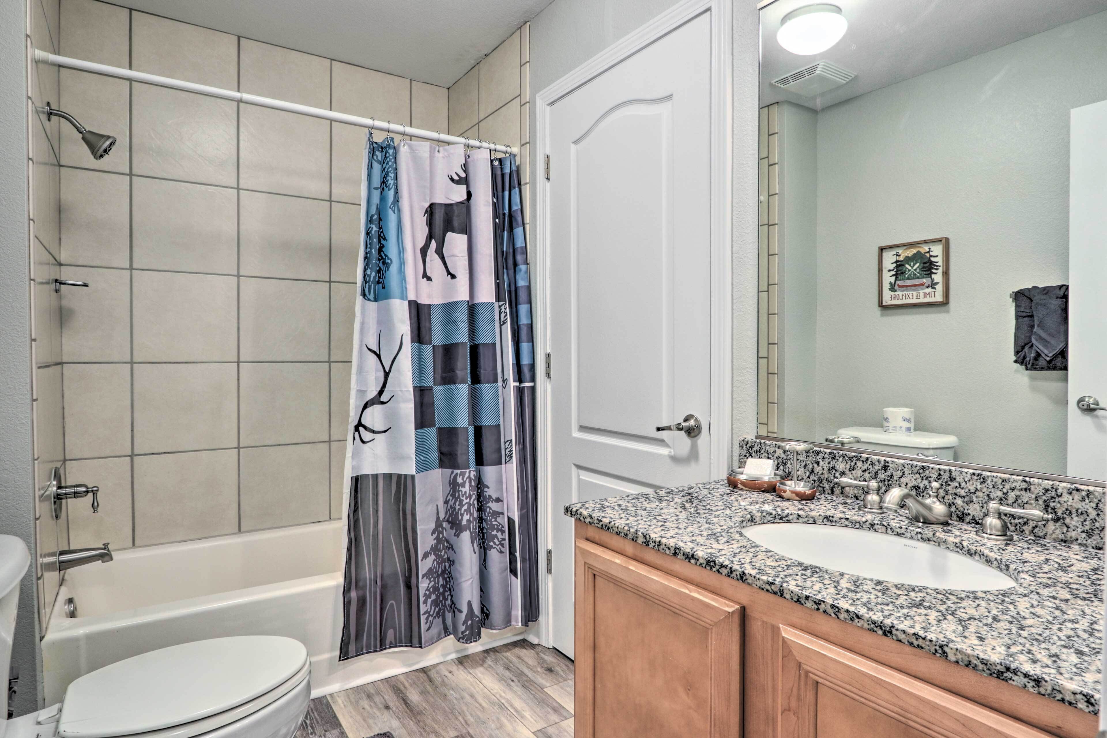 Une salle de bain avec un rideau de douche orné d’un motif représentant un cerf et un arbre, des toilettes blanches, un lavabo encastré dans un plan de travail en granit et un meuble-lavabo en bois.