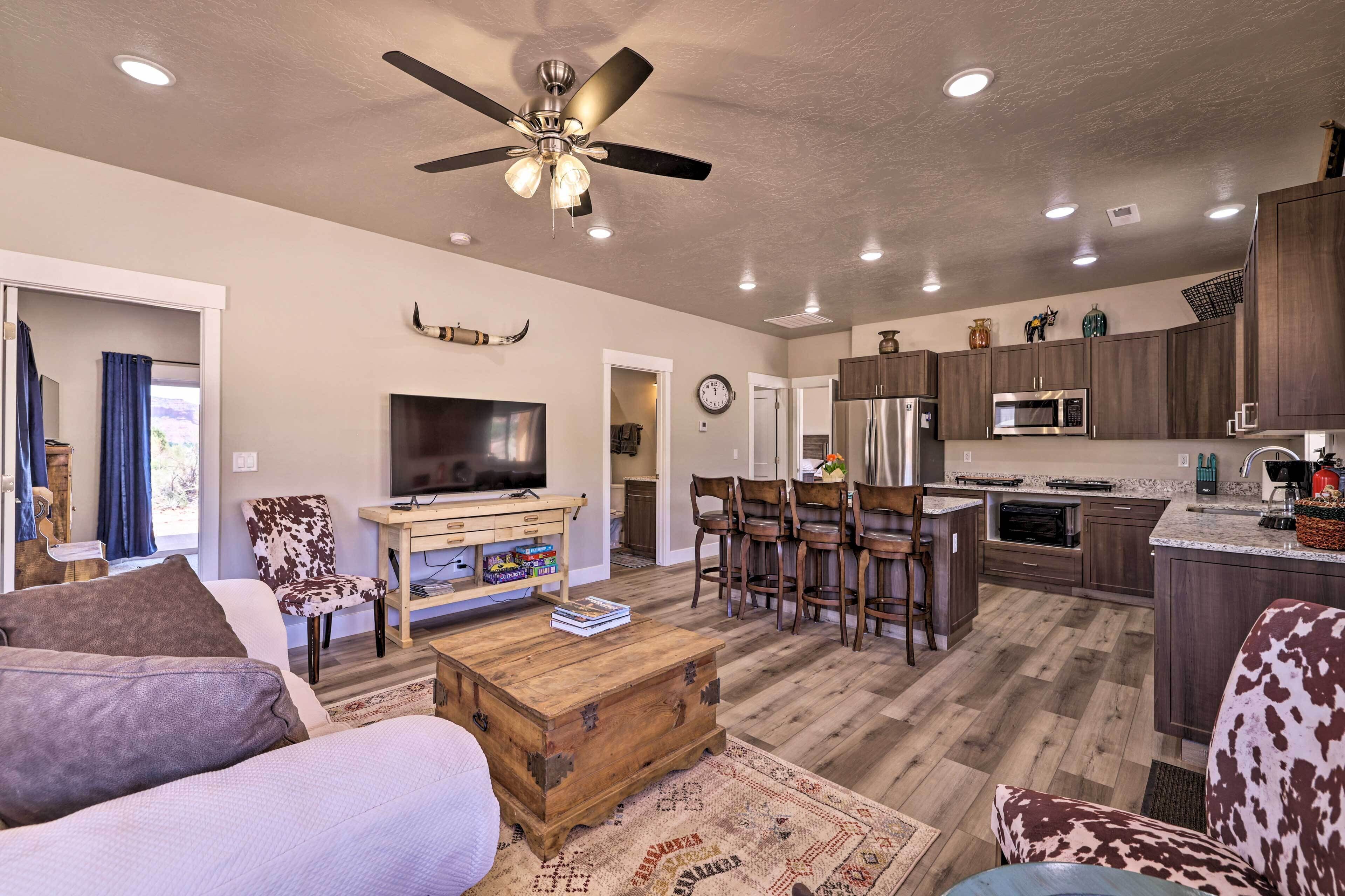 A modern living room with a sofa, coffee table, and TV. It includes a kitchen with wooden cabinets, a microwave, and a refrigerator.