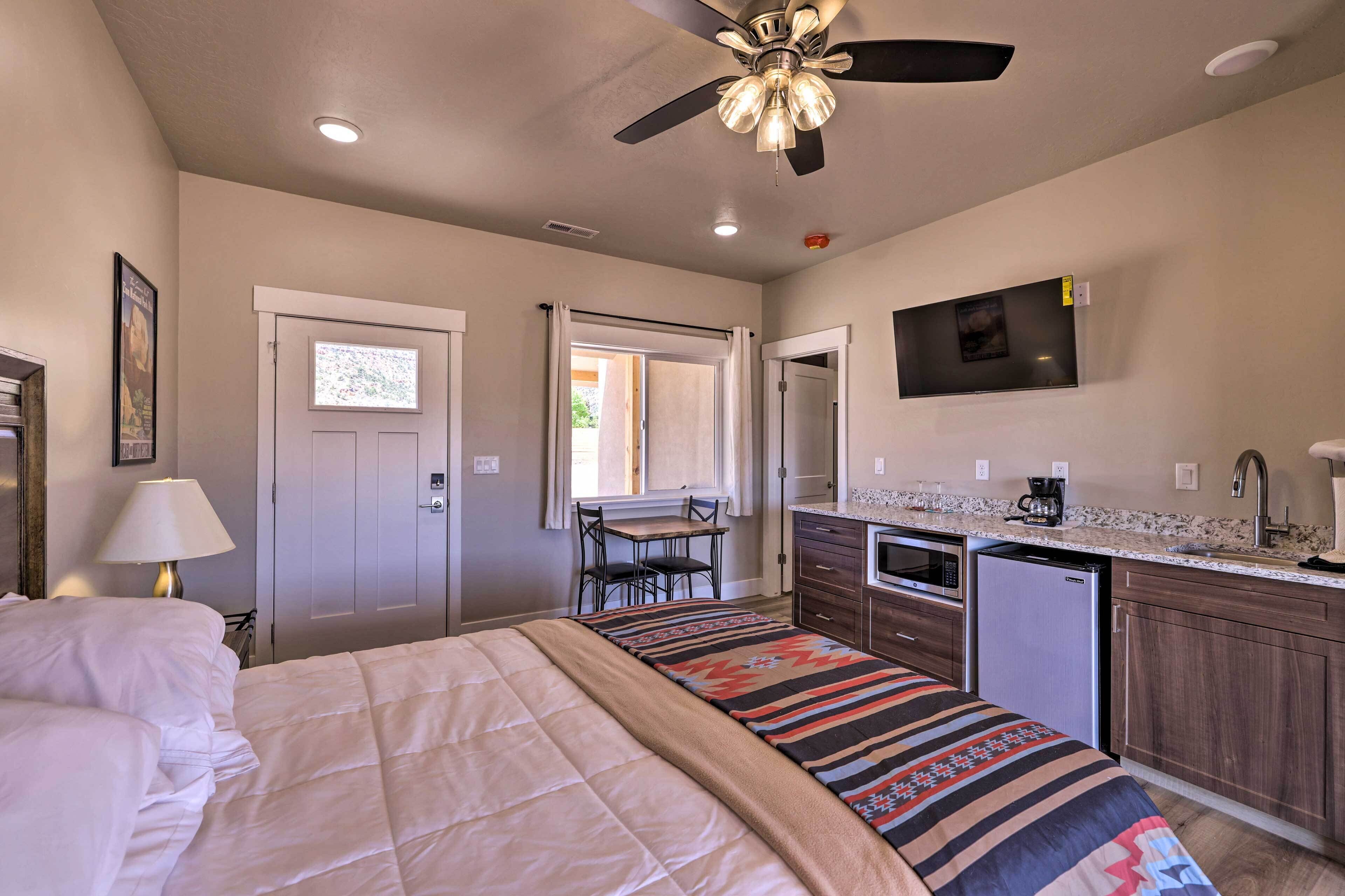 A bedroom with a bed, a desk, and a chair. There is a kitchenette with a refrigerator, microwave, and coffee maker. A ceiling fan is visible.