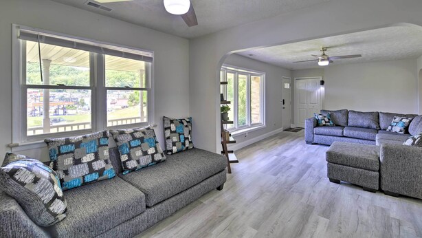 A spacious living room with a grey sectional sofa, a grey ottoman, and a grey couch. There are two ceiling fans, large windows with white frames, and a view of greenery outside. - Bluefield Retreat w/ Pool Table: Near Parks! (Bluefield)