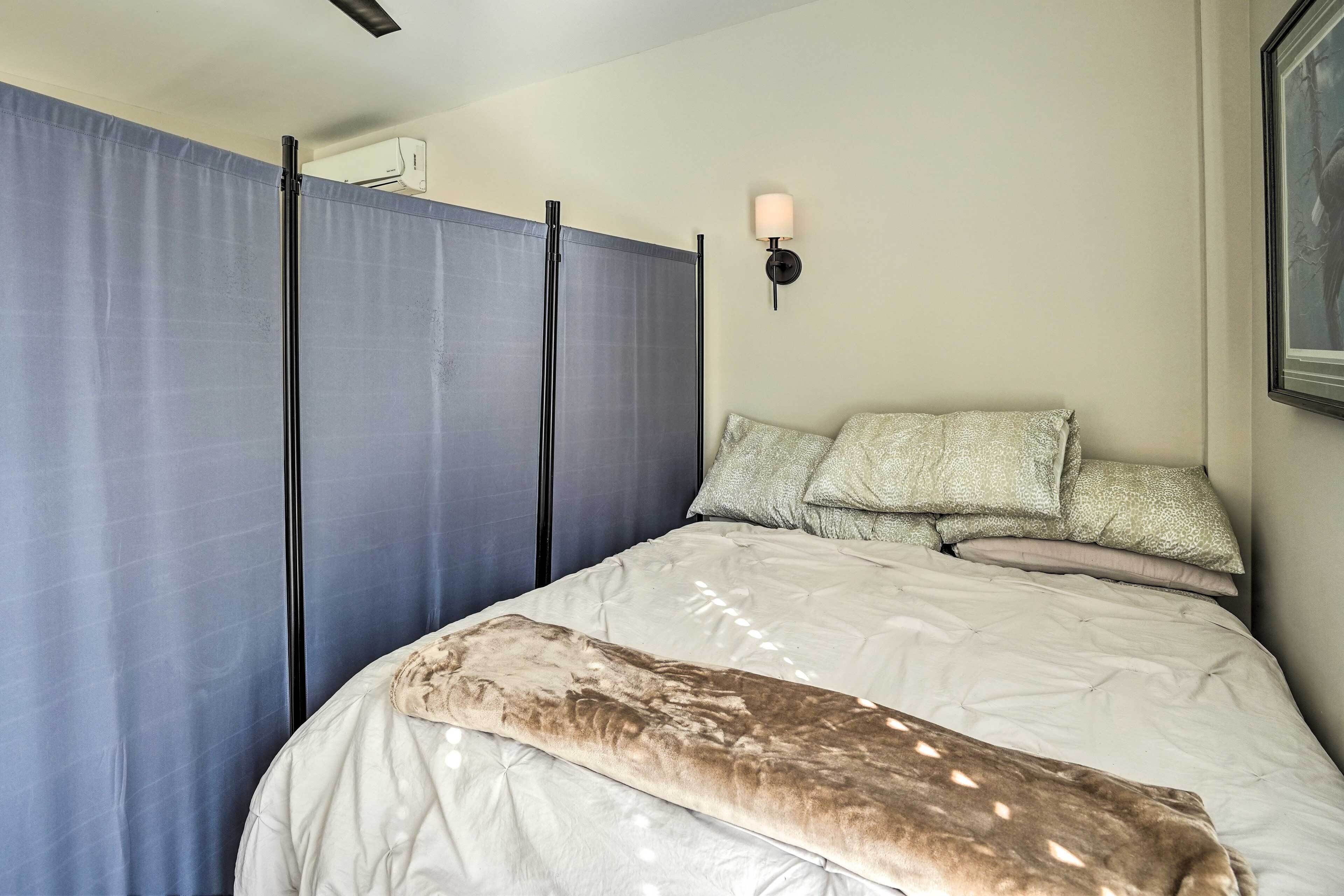 Une chambre à coucher avec un lit, des oreillers, une couverture, une lampe fixée au mur et une fenêtre avec des rideaux.
