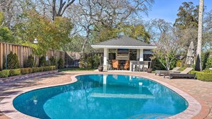Une piscine bordée de briques, un patio couvert avec des sièges et une clôture en bois en arrière-plan.