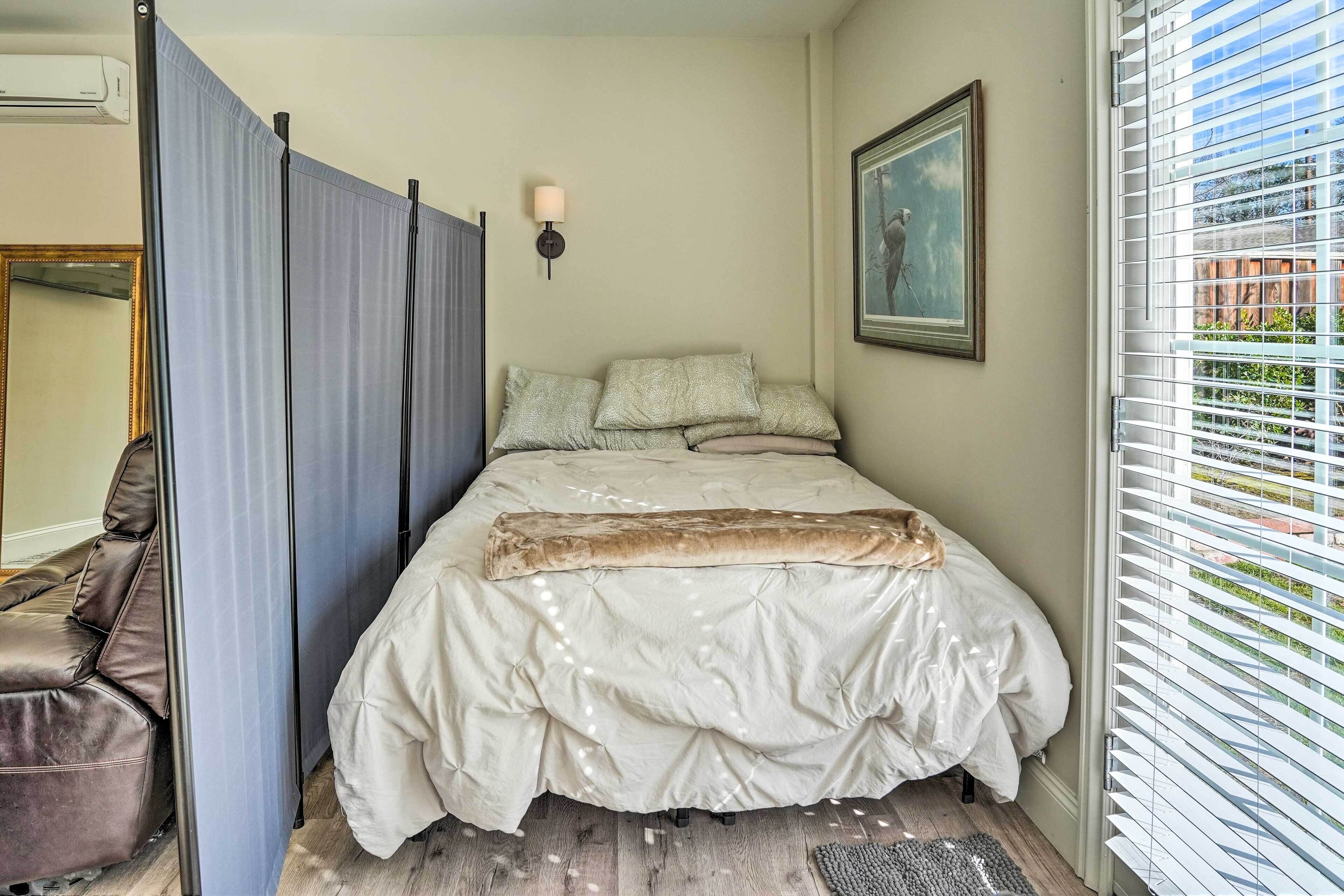 A bedroom with a bed, a window with blinds, a wall-mounted lamp, and a painting.