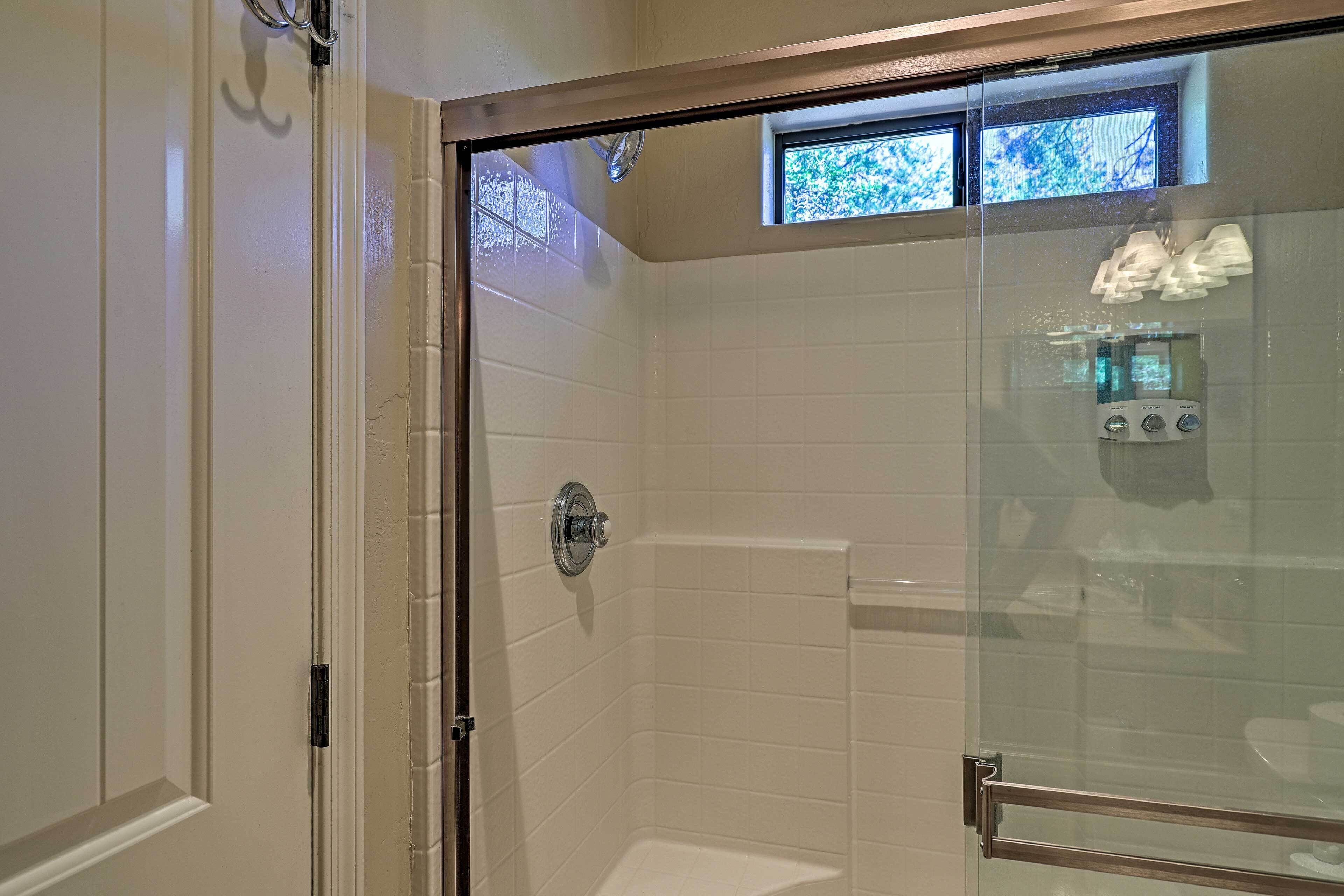 Une douche fermée par des parois de verre, avec des carreaux blancs, un panneau de commande visible et une petite fenêtre en haut.
