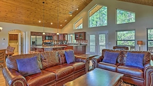 A spacious living room with leather sofas and a coffee table, a kitchen with modern appliances, and large windows offering views of the outdoors. - Serene Lakeside Cabin w/ Decks + Edler Lake Views! (Pinetop)