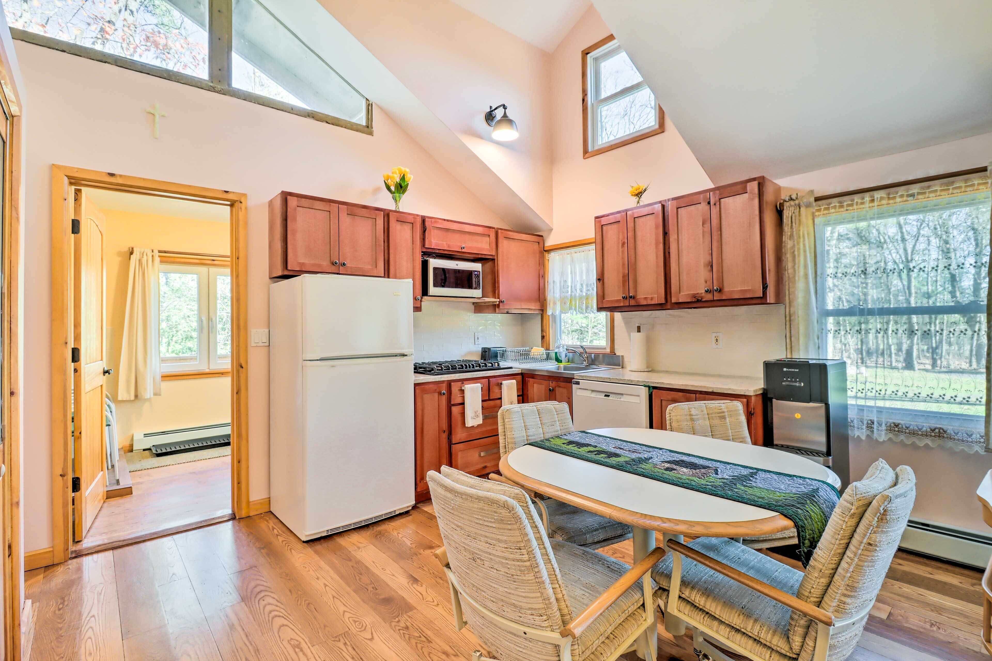 A kitchen with wooden cabinets, a refrigerator, a microwave, a stove, and a water dispenser. There is a dining table with chairs and a window with curtains.
