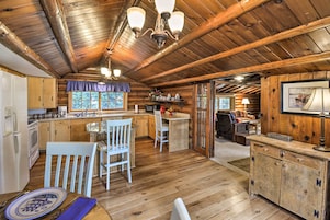 Interior rústico de cabaña con techo de madera, comedor con mesa y sillas, cocina con armarios de madera y acogedor salón con chimenea.