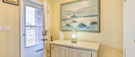 A hallway with a white door, a painting of a beach scene, and a white console table with a bottle and a vase.