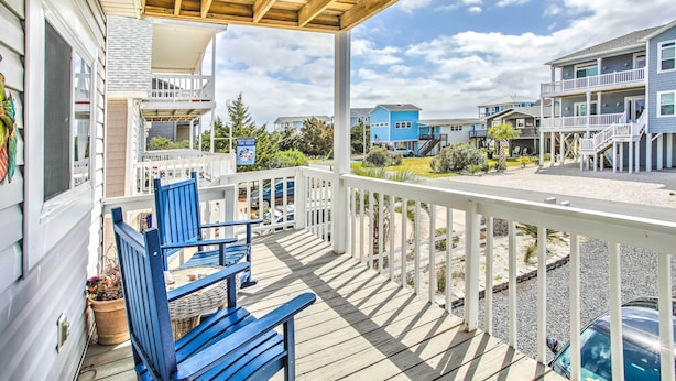 Una terraza de madera con sillas azules y una mesa, con vista a una playa y casas al fondo.