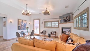 Un salon spacieux avec un grand canapé, une table à manger et deux chaises. On y trouve deux lustres, une cheminée et une grande fenêtre avec des stores.