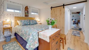 Un dormitorio con colcha estampada, cómoda de madera, mesita de noche con l ámpara y ventana grande con persianas.