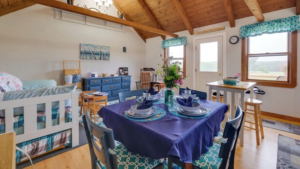 Una habitación acogedora con techo alto, vigas de madera, una mesa de comedor dispuesta con vajilla azul y blanca, una cama con colcha a cuadros azules y blancos y una ventana con vistas al exterior.