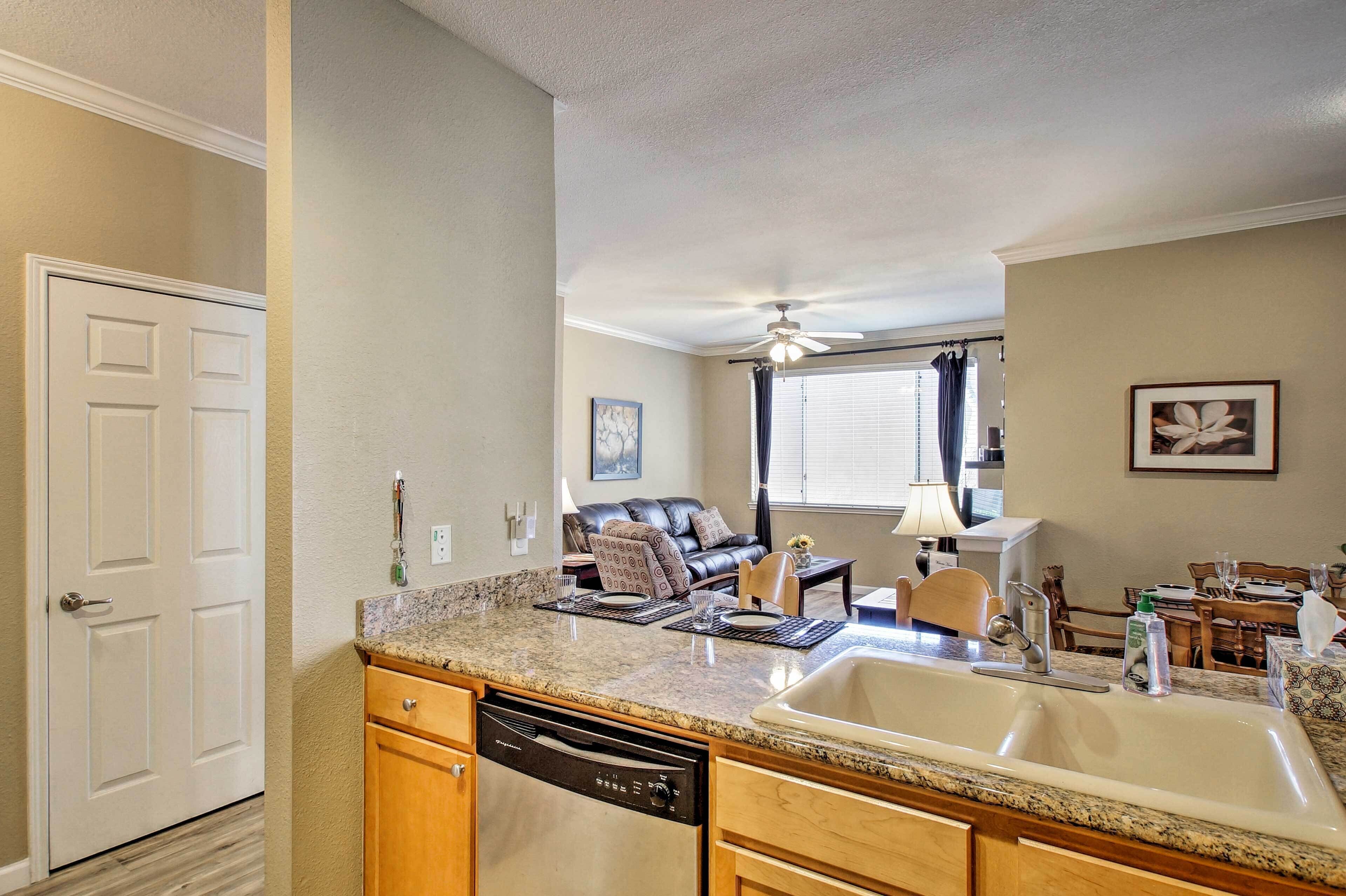 A compact kitchen with a dishwasher, granite countertop, and a sink. There is a dining area with a table and chairs, and a living room with a sofa and a ceiling fan.