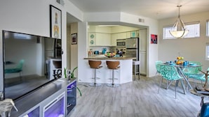A modern kitchen with white cabinets, a central island with stools, a dining area with green chairs, and a TV on a stand. - Modern Condo w/ Pool Access ~ 3 Mi to Dtwn Phoenix (Phoenix)